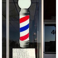 Barber shop window with a window decal of a barber shop pole and sign with text, "CLOSED UNTIL STAY AT HOME IS OVER"