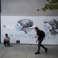 One man walking past a mural of two women wearing masks surrounded by flying birds. Another man crouches next to the mural on his phone. 