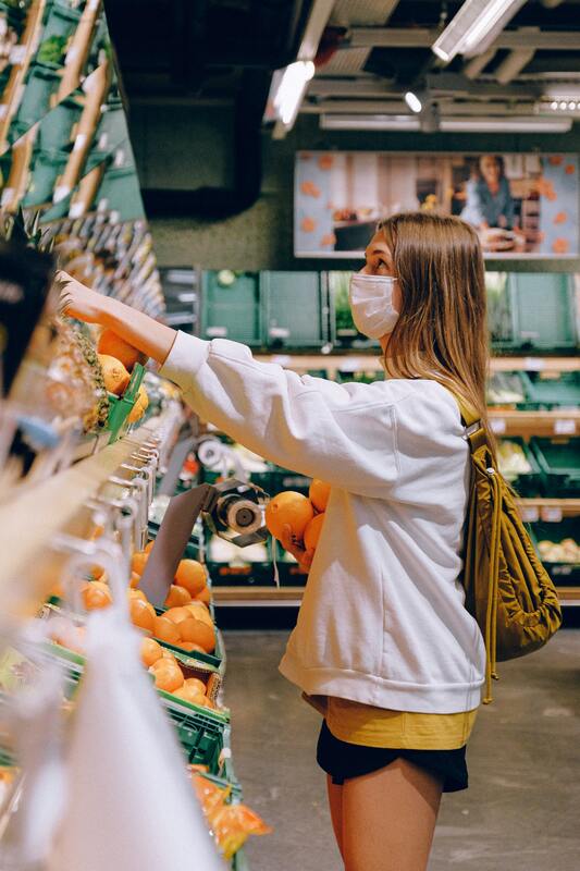 This is a picture taken of a woman who is shopping at a grocery store. She is wearing a face mask, a grey hoodie, and shorts while carrying a purse. 