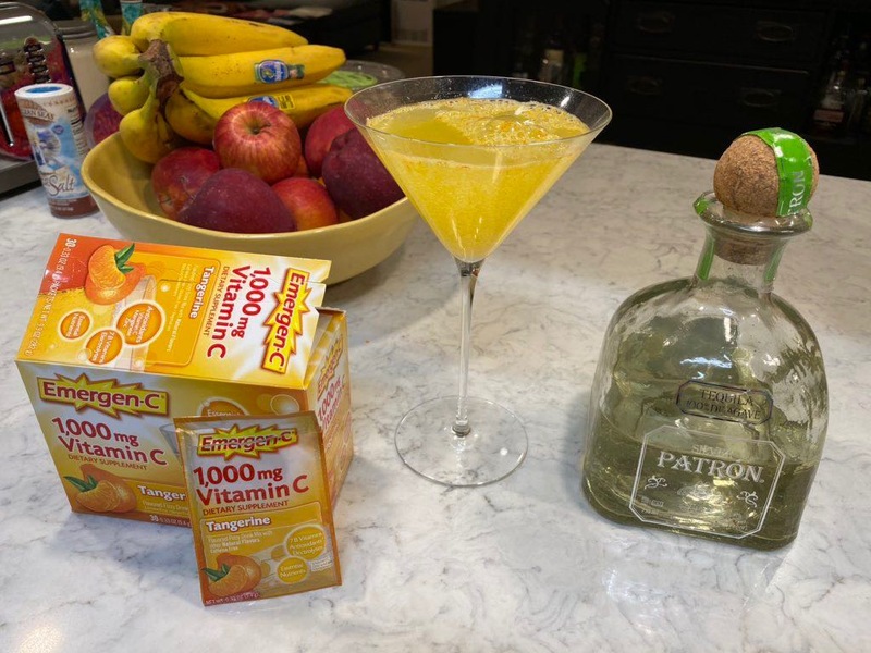 This is a picture taken of a kitchen counter, on which can be seen a bowl of fruit, a bottle of Patron tequila, a package of Emergen-C 1000 milligram vitamin C dietary supplements, and a martini glass with Emergen-C and Patron combined inside of it. 