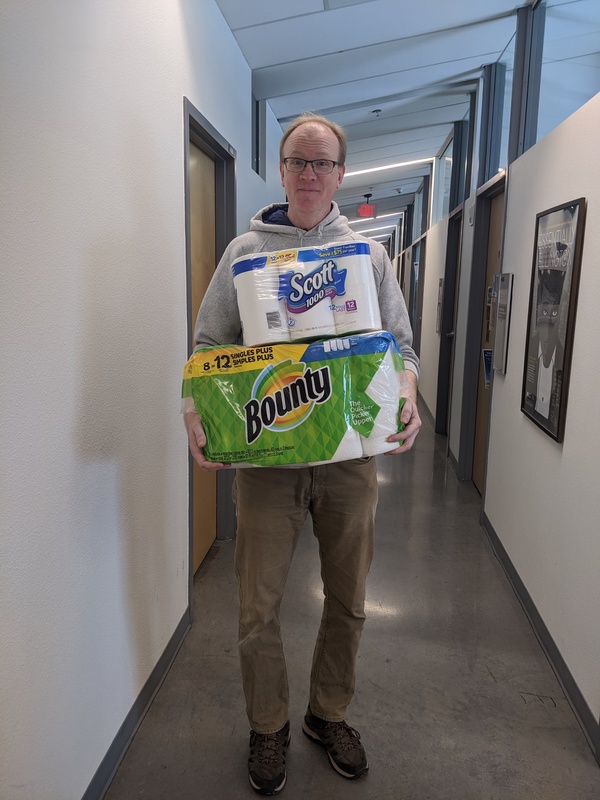 A man wearing glasses and a grey sweatshirt with khaki pants is holding a package of 12 rolls of toilet paper and 12 rolls of paper towels. 