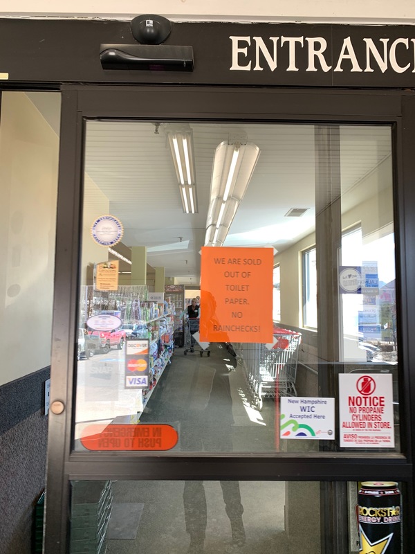 An entrance that has a sliding glass door with an orange paper sign taped on the inside of the glass that says: WE ARE OUT OF TOILET PAPER. NO RAINCHECKS!