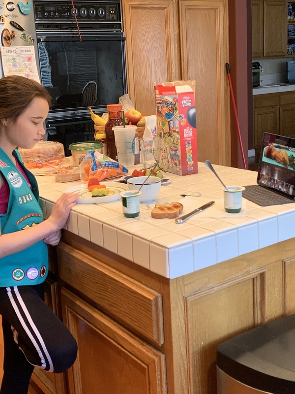 Girl in Girl Scout vest stands at a kitchen island during a virtual Girl Scout meeting. 