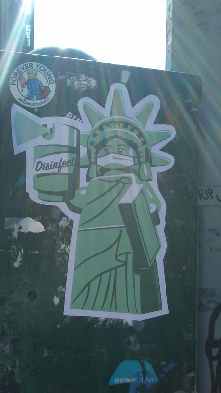 A picture of a sticker which depicts the Statue of Liberty in Lego form spraying a can of disinfectant and wearing a mask. 