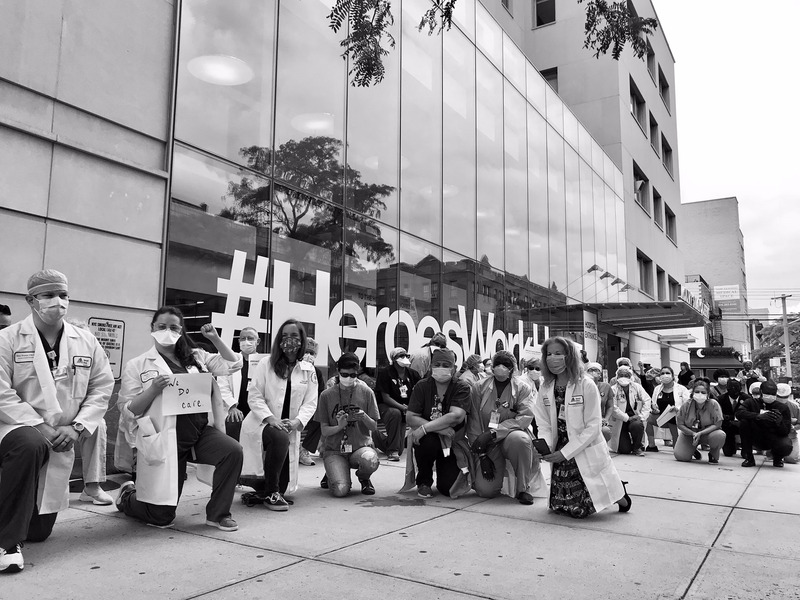 This is a picture of a group of doctors kneeling on the ground in solidarity with the Black Lives Matter movement. A large glass building is being them. 