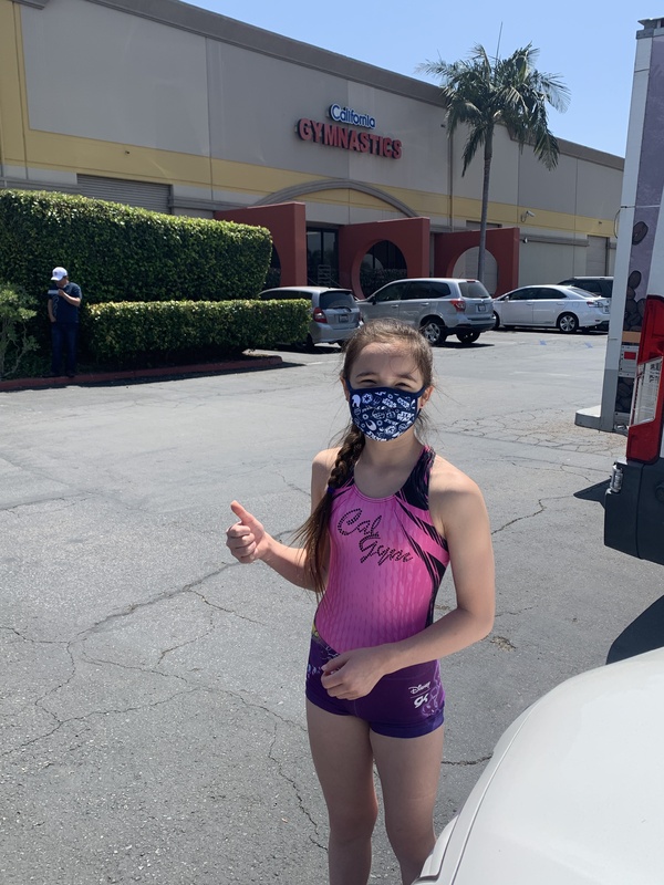 This is a picture of a young girl wearing a gymnastics uniform and a face mask, standing in front of a gymnastics building. She is giving a thumbs up sign.