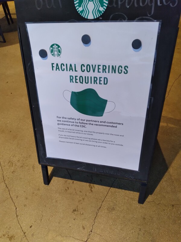 This is a picture of a sign that has been placed outside of a Starbucks coffee shop. The sign reads: "Facial covering required. For the safety of our partners and customers, we continue to follow the recommended guidance of the CDC." The image of a green face mask is in the middle of the sign. 