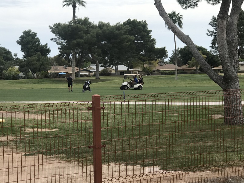A golf course that has someone golfing while two people look on while sitting in a white golf cart. 
