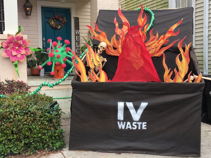 This is a picture taken of a house decorated in items related to COVID-19. A depiction of a COVID-19 germ sits on the front steps, and a fake dumpster with a skeleton and flames inside of it sits in front of the steps. The dumpster is labeled "IV Waste."