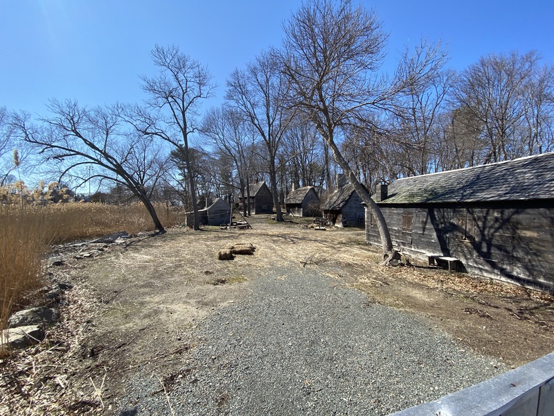 Several old worn buildings surrounded by bare trees, dry grass, and gravel at the Salem Pioneer Village in Salem, Massachusetts. 