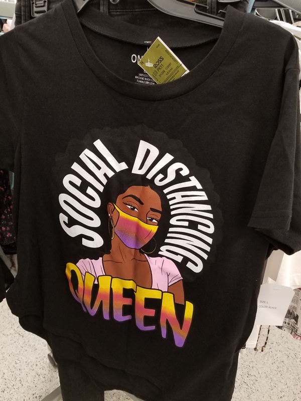 This is a picture of a black shirt which depicts a woman with an Afro wearing a face mask, and the words "Social distancing Queen" written on it. 