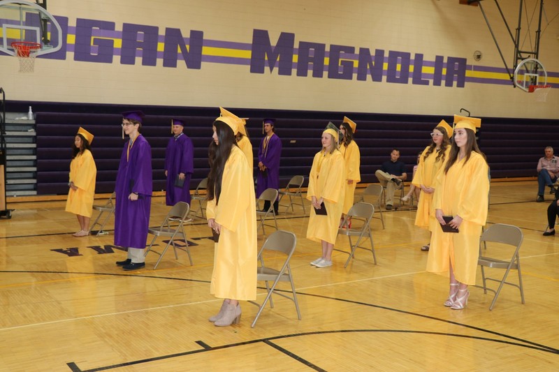 Image of soon to be high school graduates in their caps and gowns, standing spread out in a gym.