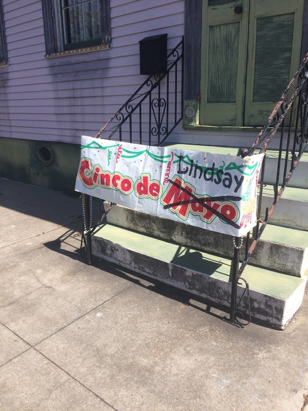 Sign attached to the railings in front of the steps leading up to double doors with text, "Cinco de Mayo" but "Mayo" is crossed out and "Lindsay" is written on the sign. 