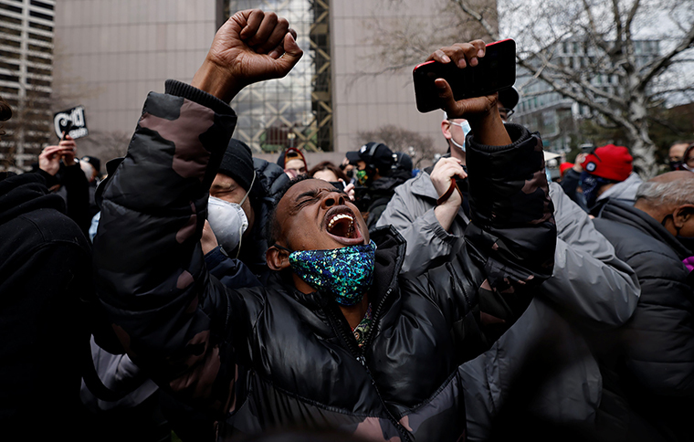 This is a picture taken of a man in a crowd who is wearing a dark winter coat, and screaming at the sky. He is holding a cell phone in his hand, and is wearing a mask on his chin. 