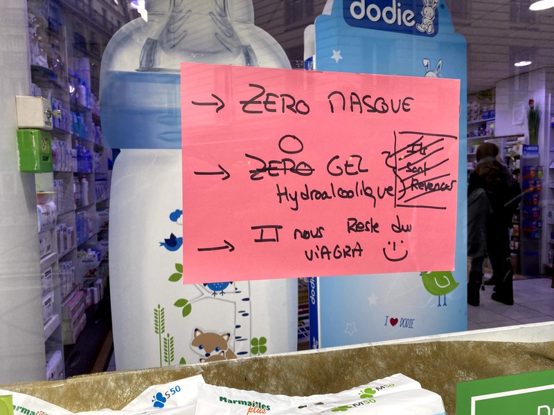 A pink piece of paper with sharpie writing on it is taped on glass window of a Pharmacy. 