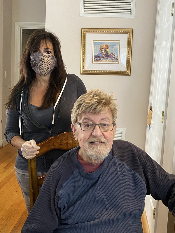 A woman wearing a cloth face mask and gloves standing behind a older man sitting in a wood chair. 