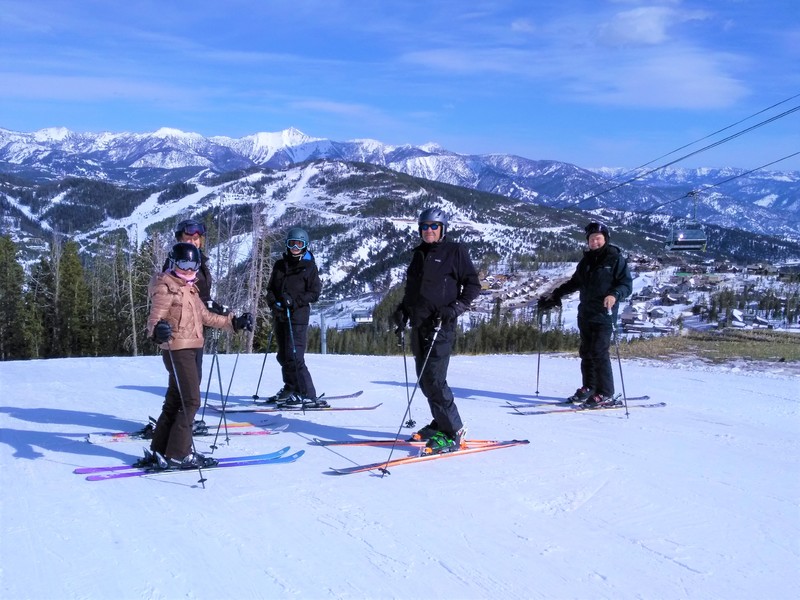 Five people posing while on skis on a snow covered hill. A gondola is going by in the background on the right. 