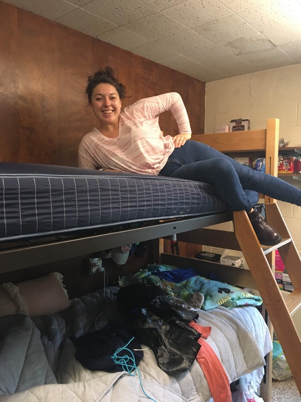 This is a picture taken of a smiling girl wearing a striped pink shirt and jeans sitting on top of the top bunk of a bunk bed. 