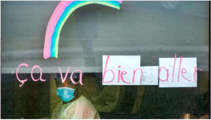 A medical worker looking out a window.  Written in window marker is the phrase "ca va bien aller" with a rainbow.