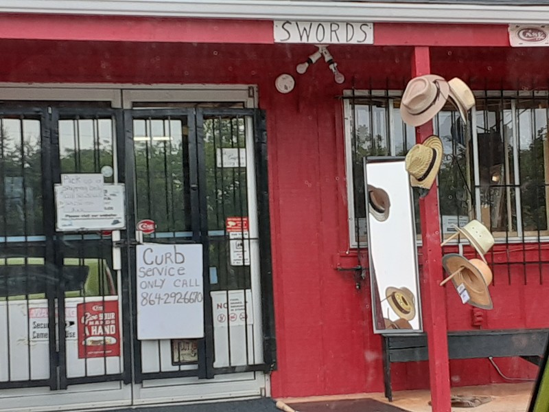 Storefront with a sign on the barred door that says "Curb service only call 846-292-6670" with multiple hats hanging in front and a sign that says "SWORDS"