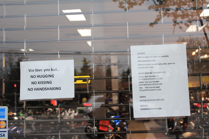 Two signs at shop.
The first sign says: "We love you but... No hugging, no kissing, no handshaking."
The second sign says: "Closed. We will reopen as soon as possible." and then gives the shop's website link along with associate's e-mail addresses and work phone numbers. 