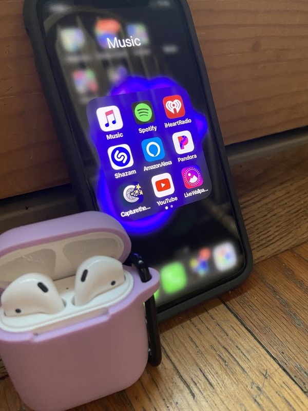 Black iPhone with music folder open. The text for apps in the music folder are, "Apple Music, Spotify, iHeartRadio, Shazam, AmazonAlexa, Pandora, Capturethe..., YouTube, LiveWallpa..." Pink case with white AirPods next on iPhone. Both items on a wood surface. 