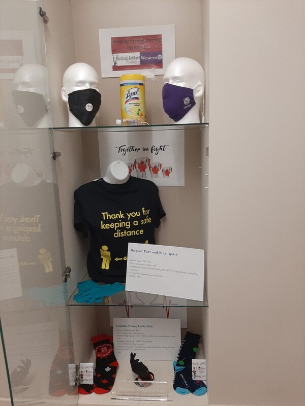 This is a picture taken of a display containing several shelf of COVID-19 related accessories. One shelf has several mannequin heads with face masks on them with a bottle of disinfectant wipes between them. A black shirt on the second shelf reads "thank you for keeping your distance." Several signs posted are encouraging the reader to "fight" against the virus, and follow social distancing standards. 