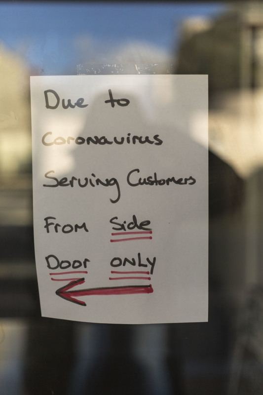 A piece of paper is taped on the inside of a window that says: Due to Coronavirus Serving customers from side door only. There is a drawn red and black left arrow on the bottom of the paper. 