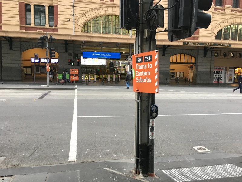 An orange sign on a stop light with directions to a tram station. Beyond the stop light is a street. 