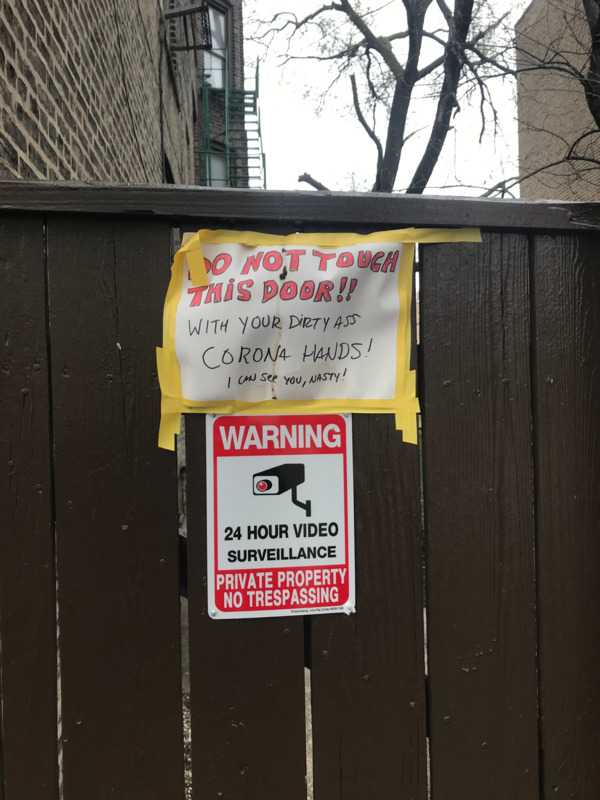 This is a picture of two paper signs taped to a wooden fence. One reads "Do not touch this door!! With your dirty ass corona hands! I can see you, nasty!". A second sign reads "Warning: 24 hour surveillance, private property, no trespassing."