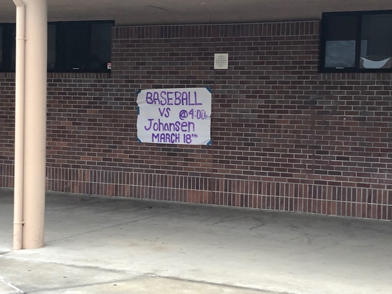 A side of a brick building. A white paper sign that is being held up by duct tape that says: BASEBALL vs @4:00pm Johansen MARCH 18th.