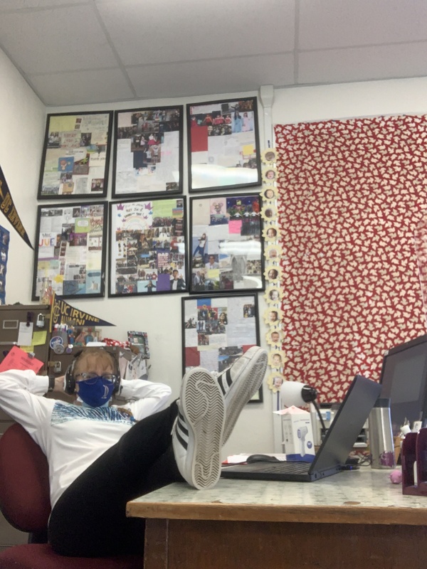This is a picture of a woman wearing a white sweatshirt, glasses, a face mask, and headphones sitting with her feet on top of a desk and her hands behind her head. She appears to be sitting in a classroom, with several collages of pictures, a computer, and various decorations set on the walls. 