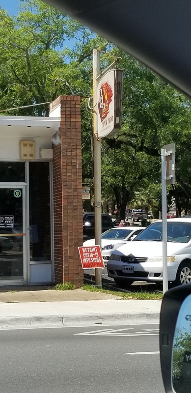 The front of a business that makes information signs. In the yard to right in front of the door has a sign that says: "We print COVID-19 info signs".
