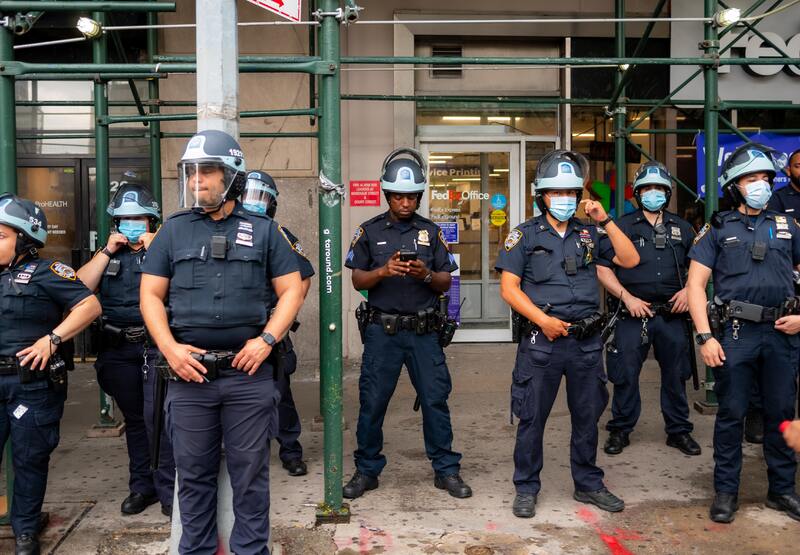 This is a picture of a group of police officers wearing both face shields and face masks standing outside of a FedEx. 