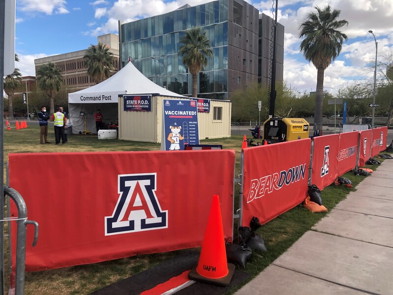 This is a picture of a vaccination command post at the University of Arizona. Several buildings and walkways are in the background, while red fencing with the U of A logo separates the command post from the sidewalk. 