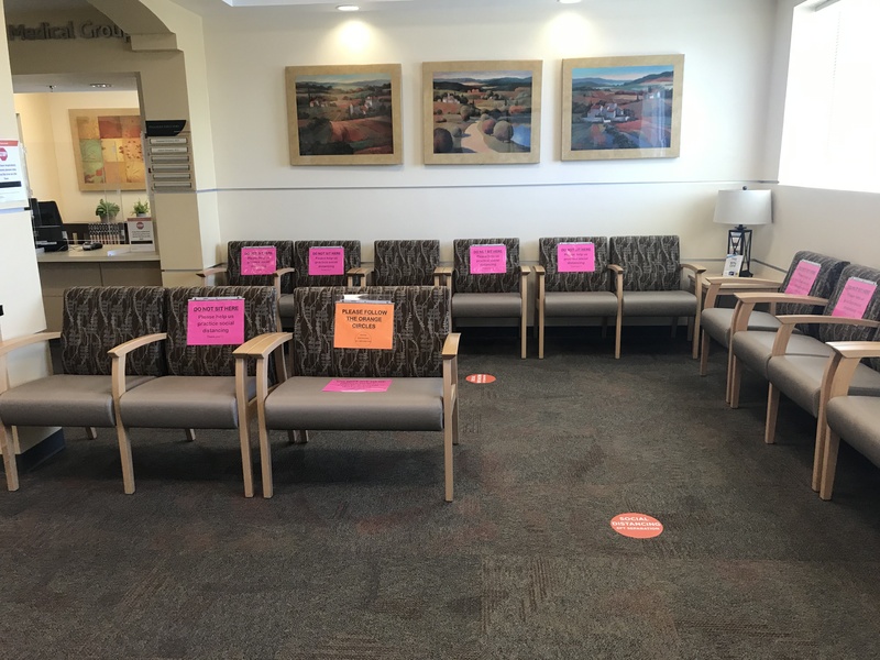 A medical center waiting room featuring chairs blocked off in order to promote social distancing. The signs on the chairs read Do Not Sit Here, please help us practice social distancing and Please Follow The Orange Circles which are on the floor indicating six feet apart for social distancing purposes. 