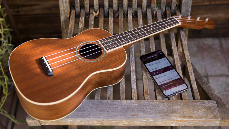 This is a picture of a four string acoustic guitar resting on top of a wooden chair next to a cell phone. 