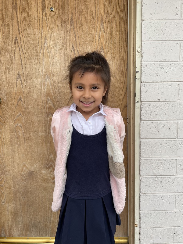 This is a picture of a girl that is standing in front of a door, smiling. She wears a fluffy pink coat, and a blue and white dress. 