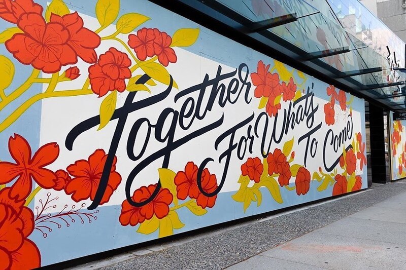 This is a picture of a mural on a wall reading "Together for what's to come" on a white and blue background, with flowers intertwined with the words. 