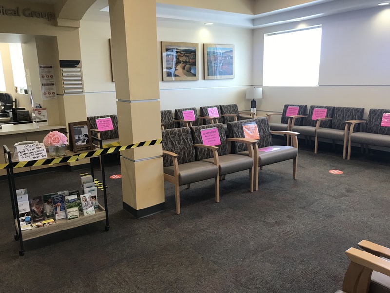 The waiting room at a medical center featuring chairs blocked off with signs saying Do Not Sit Here, Please help us practice social distancing. 