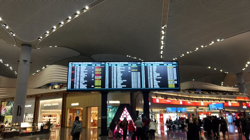 Inside of an airport that has flights on a TV screen hanging from the ceiling, most of the flights say CANCELED in yellow. The airport has people walking throughout and looking up at the TV screen for the list of flights. 