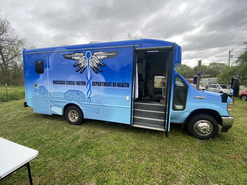 This is a picture taken of a bus parked in a grassy area. The bus is painted blue, with the words Muscogee (Creek) Nation Department of Health on the side. 