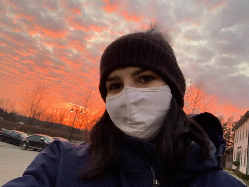 This is a picture of a woman in a blue jacket, beanie, and face mask, standing in a parking lot. 