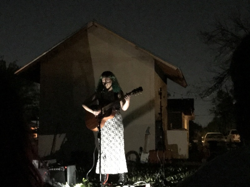 This is a picture taken of a woman who is performing music on a stage outdoors at night. She is wearing a white dress and holding a guitar, with a spotlight on her that silhouettes her picture against the wall behind her.  