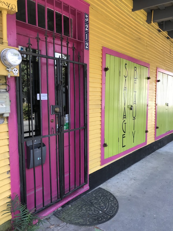The front of a bright pink, yellow, and green hair salon. The front of the bright pink doors has a black metal gate. There is a small rectangle laminated sign that is taped between the front doors. 