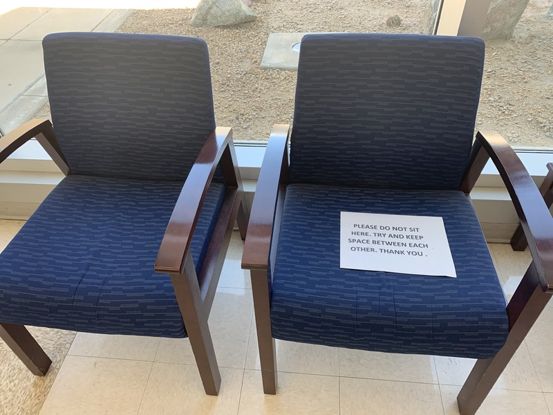Two chairs in a lobby with a paper sign on one that says: "PLEASE DO NOT SIT HERE. TRY AND KEEP SPACE BETWEEN EACH OTHER. THANK YOU."