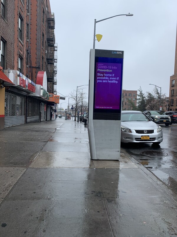 this is a picture of a 'LinkNYC" electronic advertisement kiosk on the street, which has a message on it reading: "COVID-19 Info, Prevention. Stay home if possible, even if you are healthy."