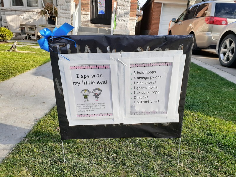 This is a picture of two signs that have been posted in front of a persons house. The first one reads "I Spy With My Little Eye! Use your spying eye to try and find the following list of items hidden in our front yard. Good luck and have fun!". A second sign reads: "3 hula hoops, 4 orange pylons, 1 pink shovel, 1 gnome home, 1 skipping rope, 2 trucks, one butterfly net". 