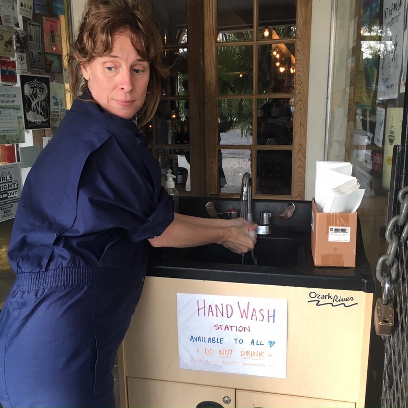 A person in navy coveralls is washing their hands at a sink. Down below in front of the cabins of the sink is a paper that says: HANDWASH STATION. AVAILABLE TO ALL. *DO NOT DRINK* 