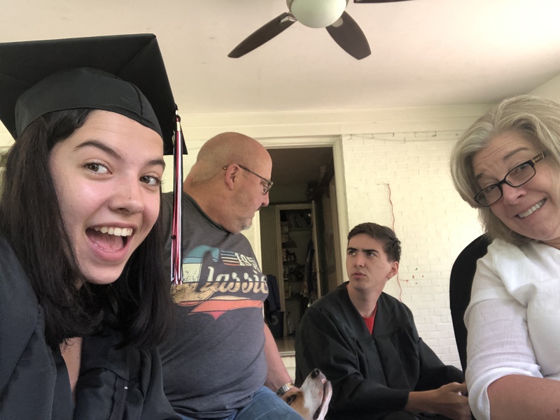 This is a picture which shows a girl and boy dressed in dark graduation clothes next to their parents in a living room. The mother and daughter are smiling into the camera, while the father and son are sharing an inquisitive look with one another. 